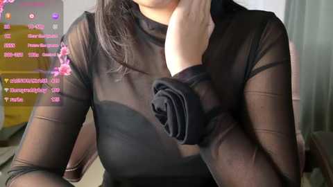 Media: Video of a woman with fair skin wearing a sheer black long-sleeve top, revealing her breast and black lace bra. She has long, dark hair and is indoors with a live stream screen in the background.