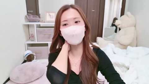 Media: A video of a young woman with long brown hair, wearing a white face mask, black top, and necklace, standing in a pastel-toned bedroom with plush toys and a white bed.