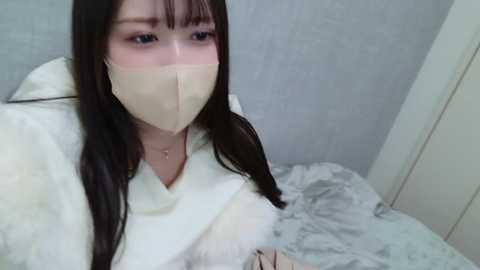 Media: Video of a young Asian woman with long black hair, wearing a beige face mask, white fur-lined coat, and grey floral-patterned bedding, with a light grey wall background.