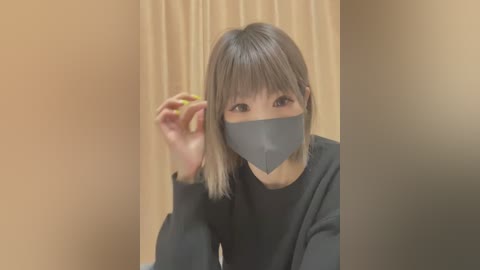 Media: Video of an East Asian woman with straight, shoulder-length gray hair, wearing a black mask, and holding a green apple in her right hand. She is dressed in a black sweater, set against a beige curtain backdrop.