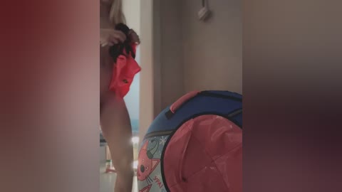 Media: A video of a nude woman with light skin and medium-sized breasts, partially visible from behind, holding a red shirt, standing next to a pink and blue inflatable pool toy in a dimly lit hallway.