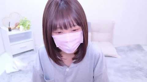 Media: Video of an Asian woman with straight brown hair, wearing a light blue shirt, pink surgical mask, and seated on a gray blanket in a white room.