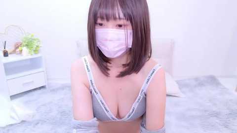 Media: A video of a young East Asian woman with straight black hair, wearing a face mask, gray bra, and a light pink shirt, kneeling in a bathtub with white walls and a white dresser in the background.