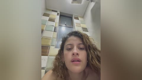 Media: A video of a young woman with curly brown hair, light skin, and full lips, standing in a bathroom with a tiled wall and a closed door.