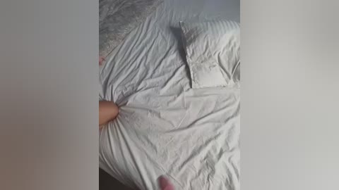 Media: A video showing a close-up of a woman's hand pulling back the corner of a white bedsheet, revealing a pillow. The room is dimly lit, with a beige wall in the background.