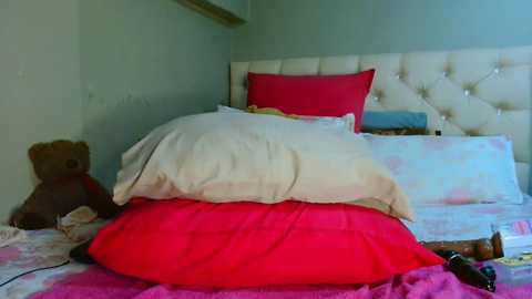 Media: Video of a neatly made bed with a tufted white headboard, featuring a teddy bear, a beige pillow, and a red pillow atop a pink duvet.