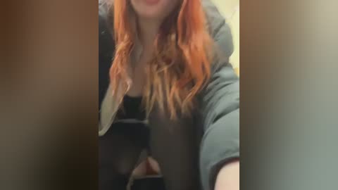 Media: Video of a young woman with long, wavy red hair, fair skin, and wearing a dark t-shirt, captured in a selfie. The background is blurred, focusing on her face and upper body.