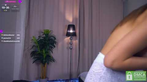 Media: A video of a nude woman with light skin, sitting on a bed, holding a towel, in a dimly lit room with a potted plant and a black lampshade.