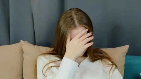 Media: Video of a young woman with long, straight brown hair, covering her face with her hand, sitting on a beige sofa with teal pillows. She wears a white long-sleeve shirt. Background features gray curtains.