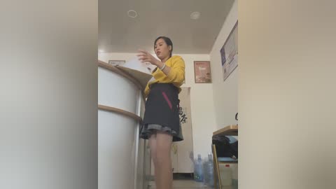 Media: Video of a woman with short black hair, wearing a yellow blouse and black skirt, gesturing in a cluttered kitchen with white walls, cabinets, and a fridge.