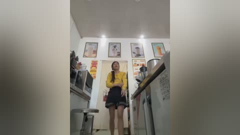 Media: Video of a young woman in a modern, brightly lit kitchen with white walls, framed art, and stainless steel appliances. She wears a yellow blouse, black skirt, and has a friendly smile.