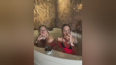 Media: Video of two women, both in bikinis, drinking from champagne flutes in a hot tub with a cave-like background. One has blonde hair, the other brunette, tattoos visible.