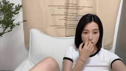 Media: Video of an Asian woman with straight black hair, wearing a white t-shirt, biting her fingernail, sitting in a white chair against a beige, text-covered curtain background.