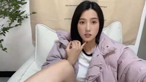 Media: Video of a young Asian woman with straight black hair, light skin, wearing a white t-shirt and lavender puffer jacket, sitting on a white chair, with a beige backdrop and a green plant in the background.