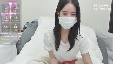 Media: A video of an East Asian woman with long black hair, wearing a white mask and T-shirt, sitting on a white bed, in a bedroom.