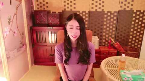 Media: Video of an Asian woman with long black hair, wearing a purple t-shirt, seated in a traditional Japanese room with wooden furniture, checkered wallpaper, and a wicker basket.