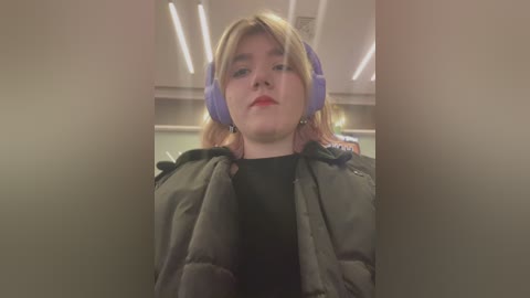 Media: Video of a young woman with blonde hair, wearing large purple headphones, a black shirt, and a puffy jacket, indoors with fluorescent lights and blurred background.