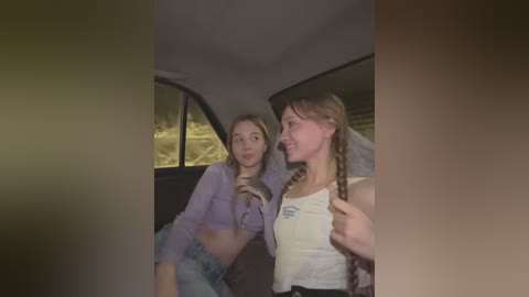 Media: Video of two young women in a dimly lit car. One with long hair in a braided ponytail, wearing a white tank top, and the other with blonde hair in a lavender crop top and jeans.