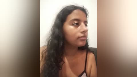 Media: Video of a young woman with long, curly black hair and medium-brown skin, wearing a black spaghetti-strap top, sitting against a plain beige wall. She has a contemplative expression.