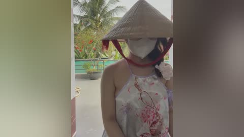 Media: Video of an Asian woman in traditional conical hat and face mask, wearing a floral-patterned sleeveless dress, standing in an open doorway with lush tropical garden in the background.