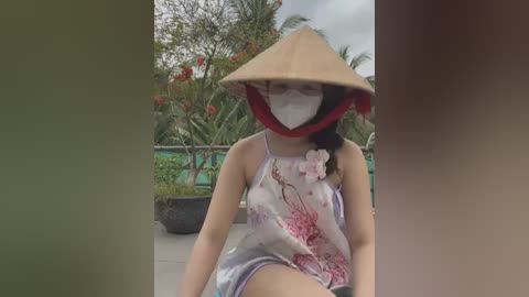 Media: Video of a woman wearing a traditional Vietnamese conical hat, floral silk blouse, and face mask, sitting outdoors with lush greenery and flowers in the background.