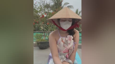 Media: Video of a young Asian woman with light skin, wearing a traditional conical hat, floral dress, and face mask, sitting on a poolside patio with greenery and red flowers in the background.