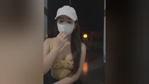 Media: Video of a young Asian woman with long black hair, wearing a white cap and mask, in a dimly lit room, holding her chin.