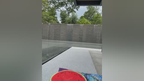 Media: Video of a minimalist outdoor pool area with a water feature, surrounded by a sleek, concrete wall with embedded vertical grooves. A vibrant, colorful watermelon slice rests on the edge of the pool.