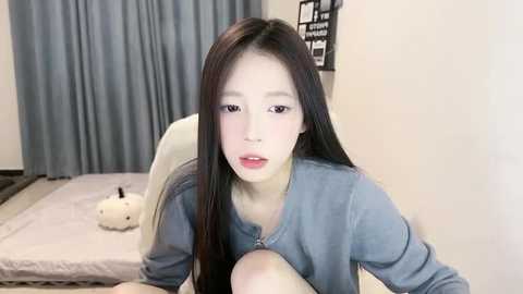 Media: Video of an East Asian woman with long black hair, fair skin, and red lipstick, wearing a blue shirt, seated on a white chair in a minimalist bedroom with light-colored walls and a gray curtain.