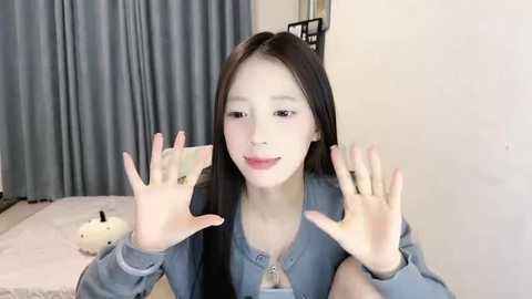 Media: Video of a young East Asian woman with long black hair, light skin, and pink lipstick, smiling and making peace signs with both hands. She wears a denim shirt. Background shows a bed with a white blanket, gray curtains, and a stuffed toy.