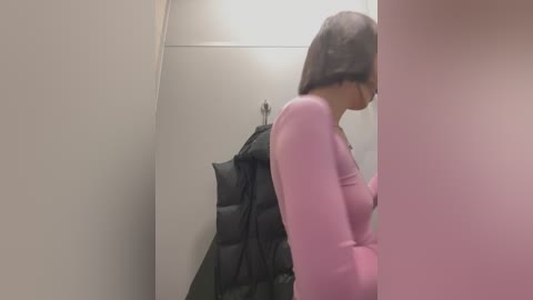 Media: Video of a woman with straight black hair, wearing a pink sweater, standing in a beige-walled changing room with a black puffer jacket hanging on a hook.