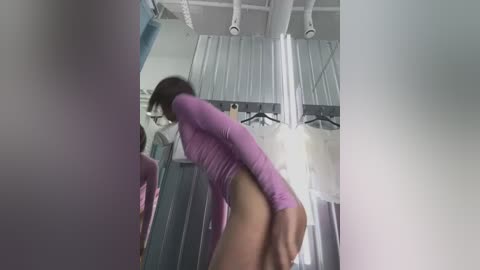 Media: Video of a woman in a lavender long-sleeve top and no pants, bending over, revealing her buttocks. She has medium-length dark hair, standing in a cluttered room with hanging clothes and a ceiling fan.