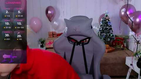 Media: Video of a cozy Christmas room with a gray cat plush toy, red balloons, a decorated tree, and a gamer in a red hoodie.