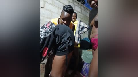 Media: Video of a young Black man with braided hair, wearing a black jacket and shorts, being restrained by a group of people, including a woman in a pink top, in a dimly lit, concrete-walled room.