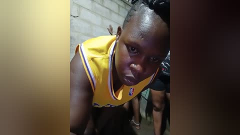 Media: Video of a young black man in a yellow basketball jersey, looking solemnly forward. He has short, tightly curled hair. Background shows a brick wall and another person partially visible.