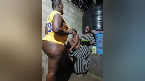 Media: Video of a stout Black woman in a yellow top and blue shorts, standing in a small, dimly lit room with two Black women in striped dresses and another man in a tank top.