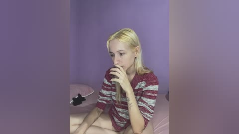 Media: Video of a blonde girl with fair skin, wearing a red and white striped sweater, sitting on a bed in a room with purple walls, holding a black object.