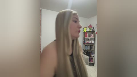 Media: Video of a fair-skinned woman with long, straight blonde hair, wearing a black sleeveless top, standing in a dimly lit room with beige walls and a bookshelf filled with various items.