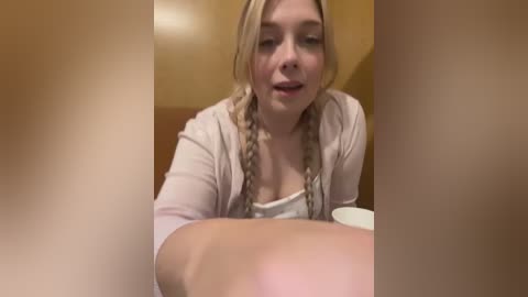 Media: Video of a young woman with blonde braids and fair skin, wearing a light pink cardigan, seated at a table. She looks slightly towards the camera with a neutral expression.