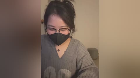 Media: Video of an East Asian woman with glasses, wearing a black face mask and grey sweater with \"LOVE\" in white, standing in a dimly lit room.