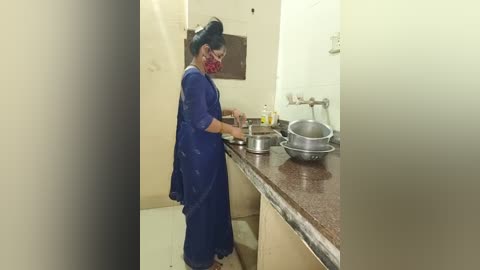 Media: Video of a woman in a blue saree with a red face mask, washing dishes in a small, sparsely furnished kitchen with a granite countertop and two metal bowls.