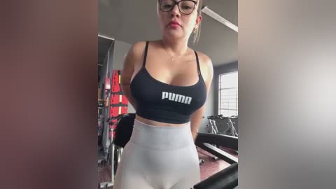 Media: Video of a fit, light-skinned woman with glasses, wearing a black Puma sports bra and white high-waisted leggings, standing in a gym with weights and equipment visible in the background.