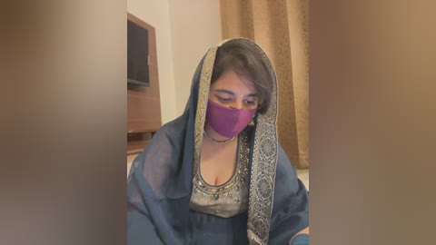 Media: Video of a woman wearing a blue, embroidered dupatta over her head, purple mask, and a silver blouse, seated indoors with a TV and beige curtains in the background.