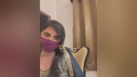 Media: A video shows a woman with long black hair, wearing a purple mask and a striped shirt, peeking from behind a doorway. She has a small handbag and beige curtains in the background.