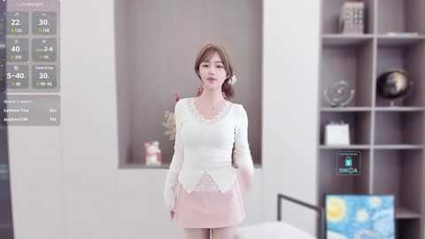 Media: Video of a young Asian woman with fair skin and straight brown hair in a white lace top, pink skirt, and headphones, standing in a modern, minimalist room with a digital display and bookshelves.