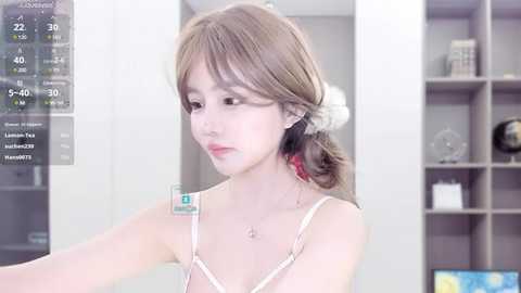 Media: Video of a young Asian woman with fair skin and straight, light brown hair in a side ponytail, wearing a white camisole and a delicate necklace. Background includes a modern, minimalist room with shelves and a clock displaying 9:40.