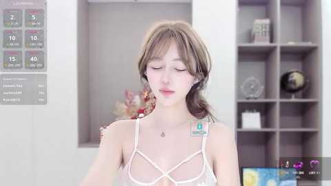 Media: Video of a young Asian woman with light skin, wearing a white lace bra, standing in a modern, minimalist room with a weather chart and shelves.