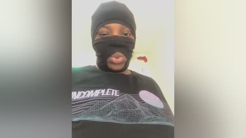 Media: Video of a Black man with a light complexion wearing a black mask, black hat, and a black shirt featuring a white graphic. He sticks out his tongue. Background is a plain, light-colored wall.