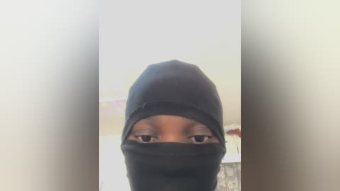 Media: A video of a person wearing a black hooded mask, revealing only their eyes, set against a blurred indoor background with a white ceiling. The image has a grainy, slightly out-of-focus quality.