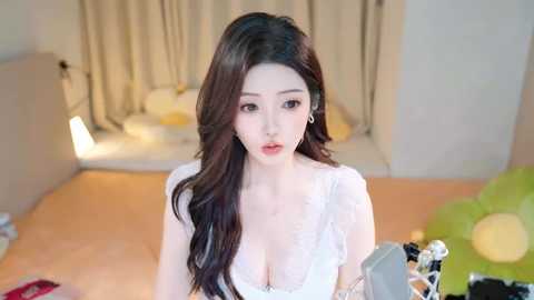 Media: Video of a young East Asian woman with long, wavy black hair, fair skin, and large eyes, wearing a white lace bra, standing in a modern, softly lit bedroom with beige curtains and a green pillow.